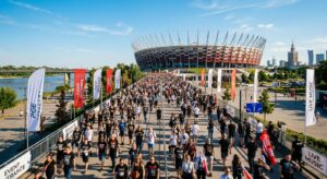 A high-angle editorial photograph of commuters walking towards Warsaw&rsquo;s National Stadium under a cle